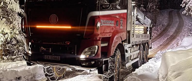 A red and white TATRA truck navigates a wintry road, flanked by a dense forest under a dark night sky.