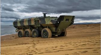 An IDV amphibious combat vehicle climbs up on the sand next to a body of water.