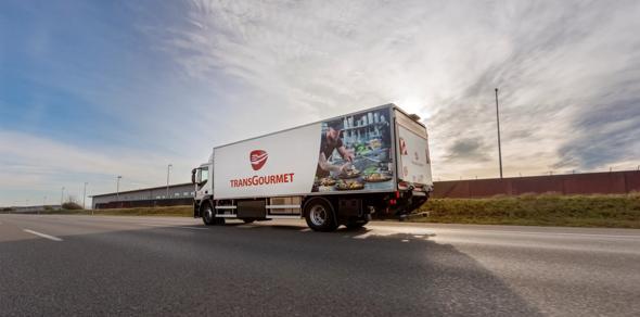 A photo of a white  TransGourmet distribution truck driving down a road.