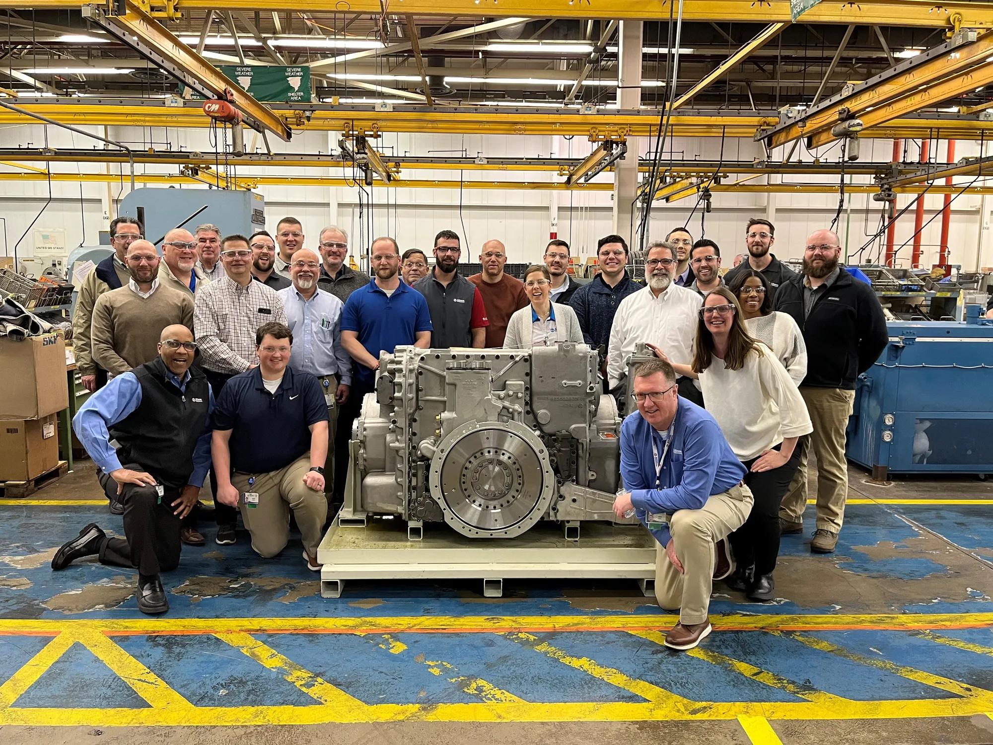 A group of people in work clothes pose for a photo in front of an X1100 transmission. Some are smiling, some are looking at the camera. They are in a factory setting. There are overhead yellow cranes with red and green safety signs. The factory floor is blue with yellow painted lines.