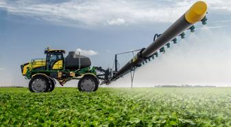 An agriculture sprayer, painted green and yellow, is applying pesticides to a field of soybeans.