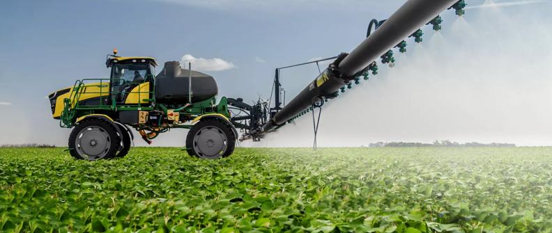 An agriculture sprayer, painted green and yellow, is applying pesticides to a field of soybeans.