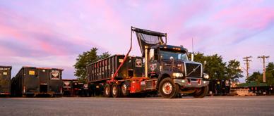 A black refuse truck hauling a dumpster with a sunset background.