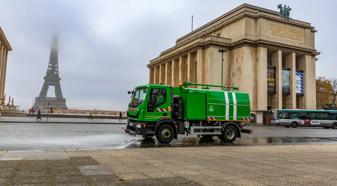 A green street cleaning vehicle sprays water on the cobblestone street in front of the Musee de l'Homme. The Eiffel Tower is faintly visible in the background through the mist.