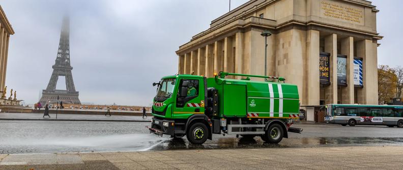 A green street cleaning vehicle sprays water on the cobblestone street in front of the Musee de l'Homme. The Eiffel Tower is faintly visible in the background through the mist.