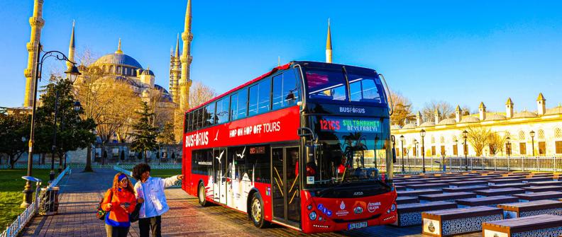 An image of a red double decker bus advertising Hop on Hop Off tours in front of the main mosque in Istanbul.