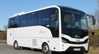 A white and black AOS coach bus with the logo "Kelacoach" on the side. The bus is parked on a paved road with a hedge on the right. The sky is blue and the sun is shining.