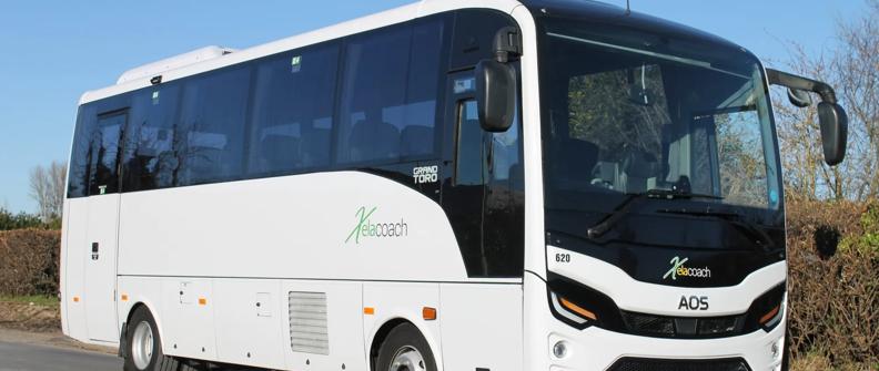 A white and black AOS coach bus with the logo "Kelacoach" on the side. The bus is parked on a paved road with a hedge on the right. The sky is blue and the sun is shining.