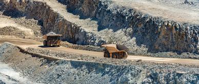 Large industrial mining trucks transport heavy copper ore along a dirt haul road cut into the rocky terraced walls of a massive open pit mine in Chile.