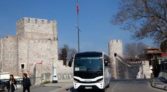 A white tour bus travels on paved road with a castle wall in the background.